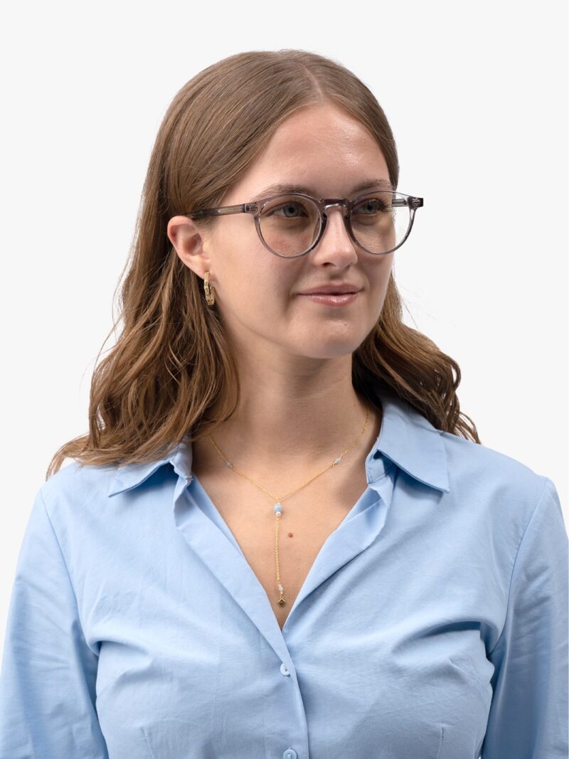 Frau mit grauer runder Brille, hellblauem Hemd, goldener Y-Kette mit blauem Anhänger und goldenen Ohrringen