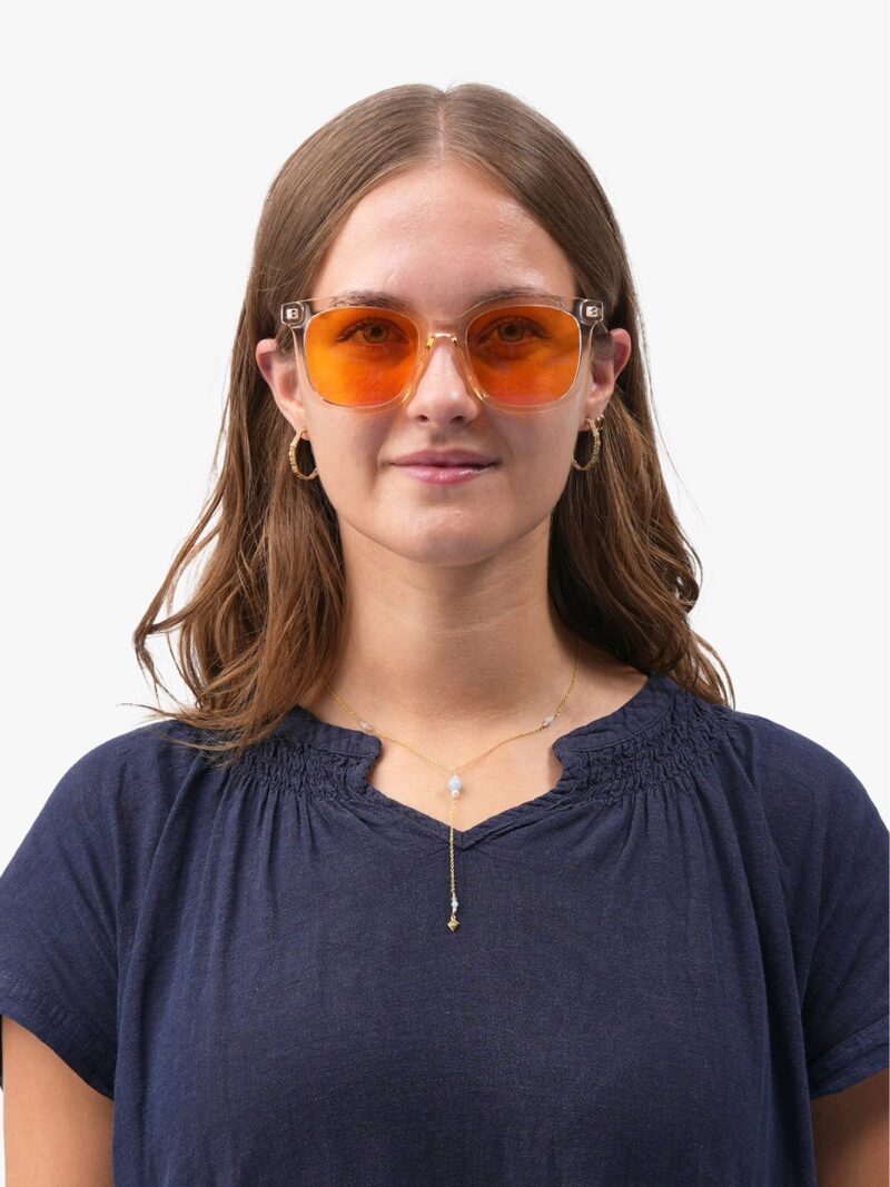 Frau mit orange getönter Sonnenbrille und transparentem Gestell, goldene Halskette und Creolen, dunkelblaue Bluse
