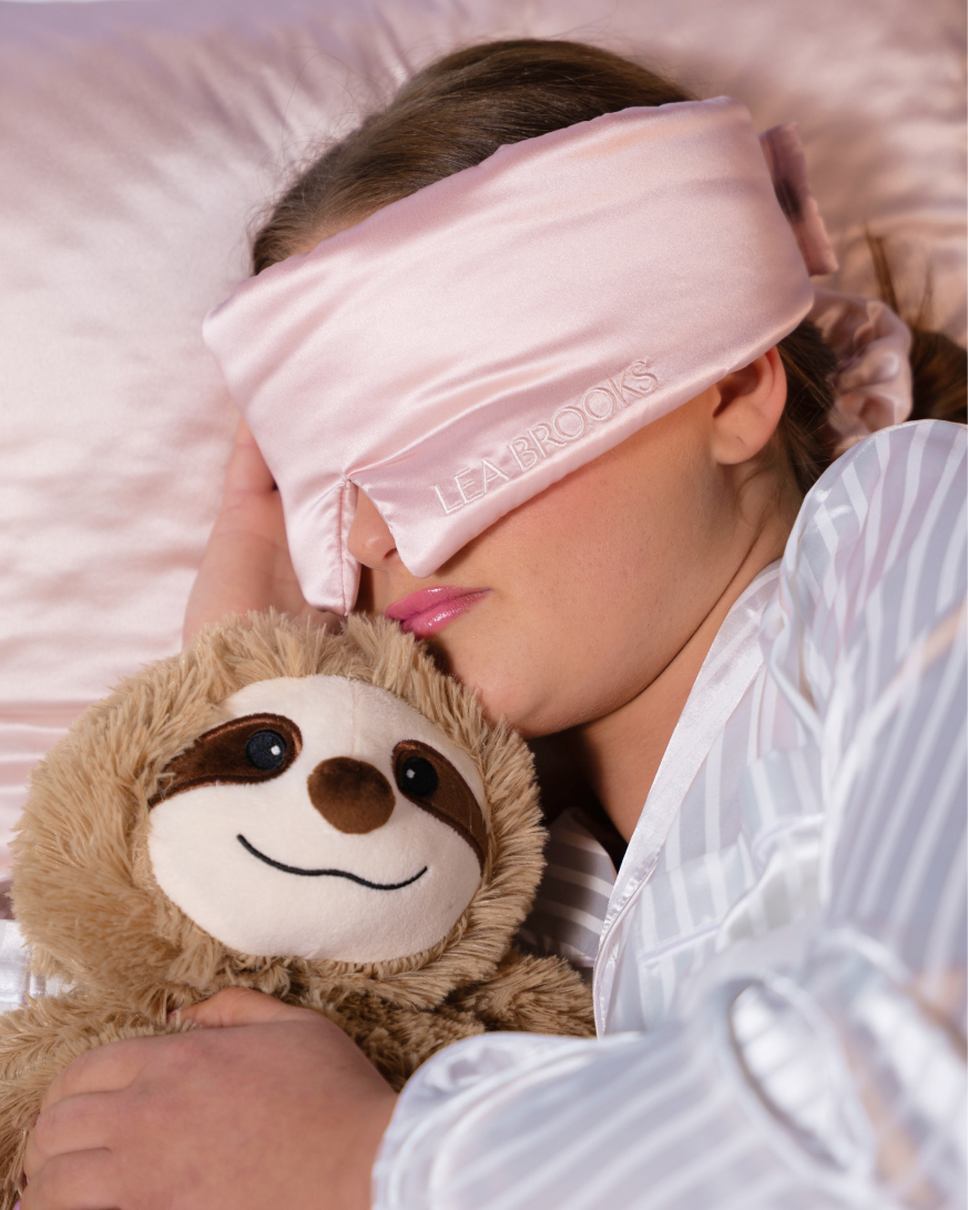 Person schläft mit rosafarbener Seiden-Schlafmaske und gestreiftem Pyjama und kuschelt mit einem weichen Faultier-Kuscheltier