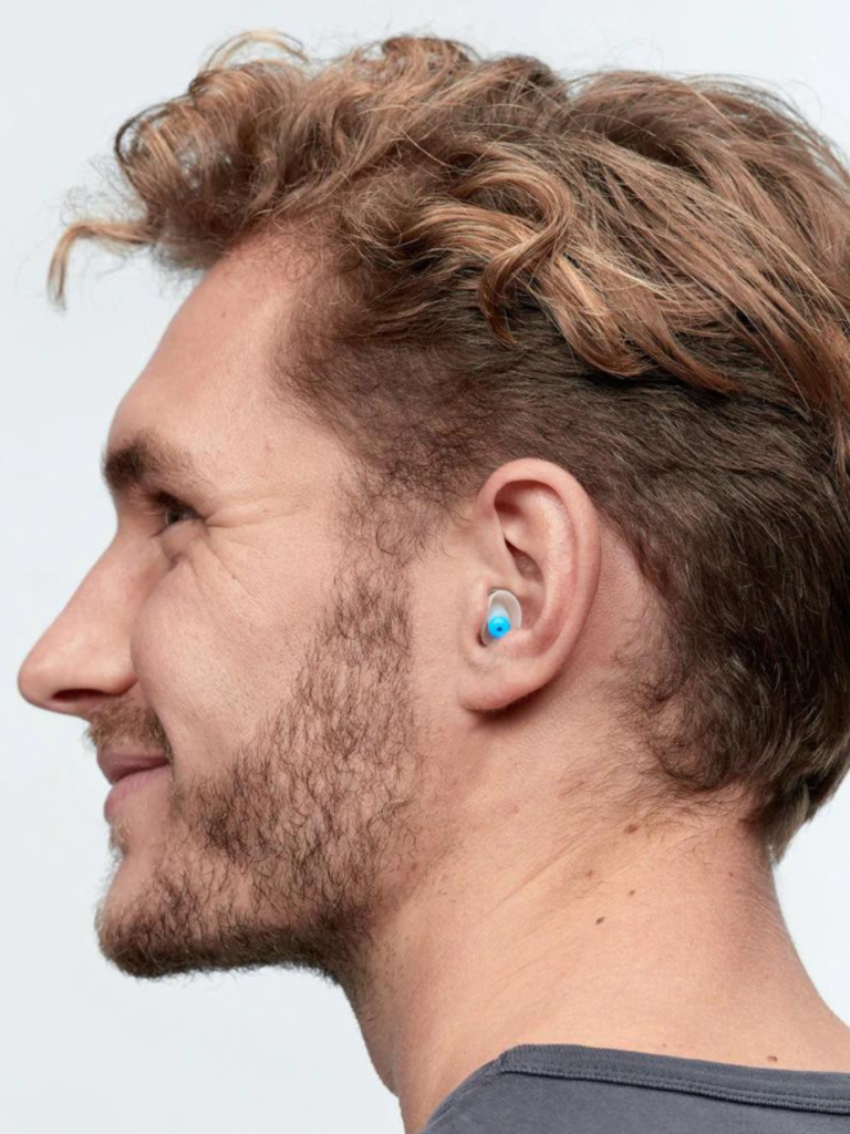 Seitenprofil eines lächelnden Mannes mit lockigem blondbraunem Haar und blauem In‑Ear‑Ohrstöpsel